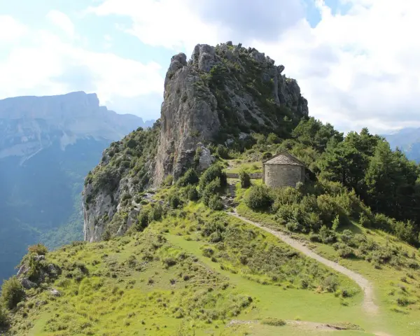 Pequeña ermita de piedra con tejado cónico, rodeada de vegetación y un sendero que sube por la ladera. Las ermitas de Tella se alzan majestuosas.
