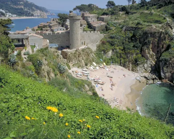 Vista de las calas de Tossa de Mar, con un castillo medieval y una playa con gente disfrutando del sol y el mar.