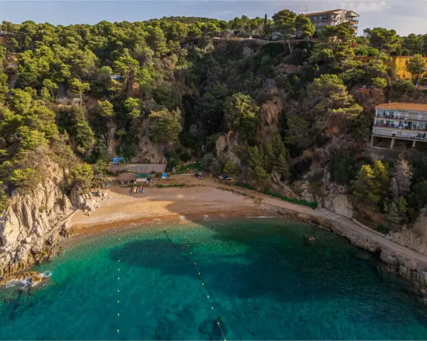 Bahía de Cala Bona, Barcelona, con aguas turquesas, arena dorada y acantilados verdes. Casas en la ladera.