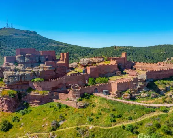 Imponente castillo de Peracense, una fortaleza de piedra rojiza que se alza sobre un peñón, rodeada de vegetación y bajo un cielo azul intenso.