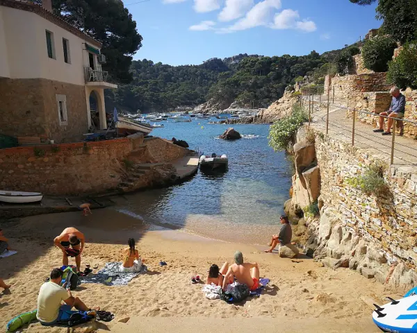 Gente relajándose en la arena de cala Malaret, Begur, con barcos anclados en aguas cristalinas y colinas verdes al fondo.