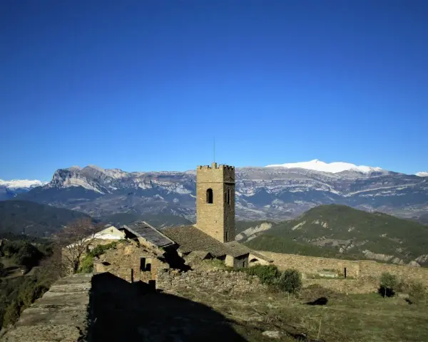 Antiguo muro de roda con torreón de piedra y ruinas, rodeado de montañas nevadas bajo un cielo azul intenso.