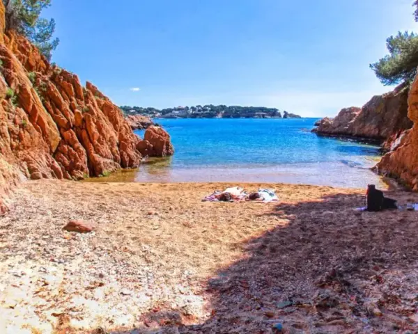 Arena dorada y aguas turquesas en la cala Maset. Dos personas toman el sol tranquilamente en la playa, rodeadas de imponentes rocas rojizas.