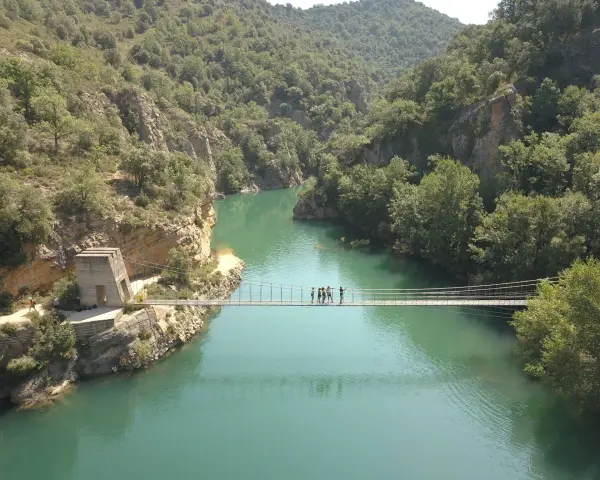 Un puente colgante cruza un río turquesa rodeado de montañas verdes. Un grupo de personas camina por el puente de Montañana.