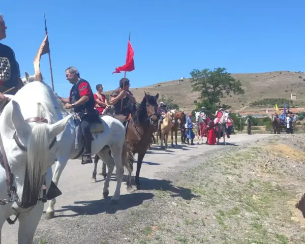 Un desfile ecuestre con trajes históricos, quizás recreando la gesta de el poyo del cid, avanza por un camino rural bajo un cielo azul.