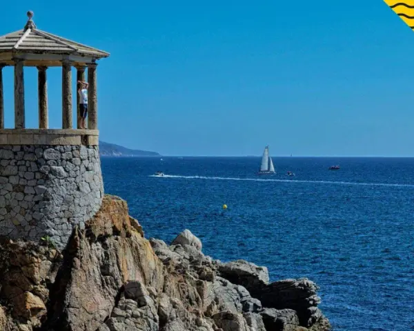 Un mirador de piedra con columnas se alza sobre rocas, con el mar azul y veleros al fondo. Un lugar perfecto para pasear por los **caminos de ronda**.