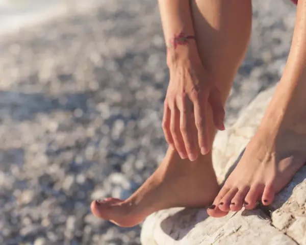 Pies descalzos sobre rocas en una playa nudista de Cadaqués. El sol brilla sobre la piel mojada y un pequeño tatuaje de flores adorna el brazo.