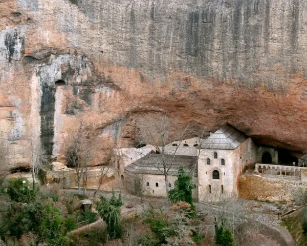 Monasterio de San Juan de la Peña, un impresionante conjunto arquitectónico excavado en la roca, con edificios de piedra y tejados a dos aguas bajo un gran saliente rocoso.