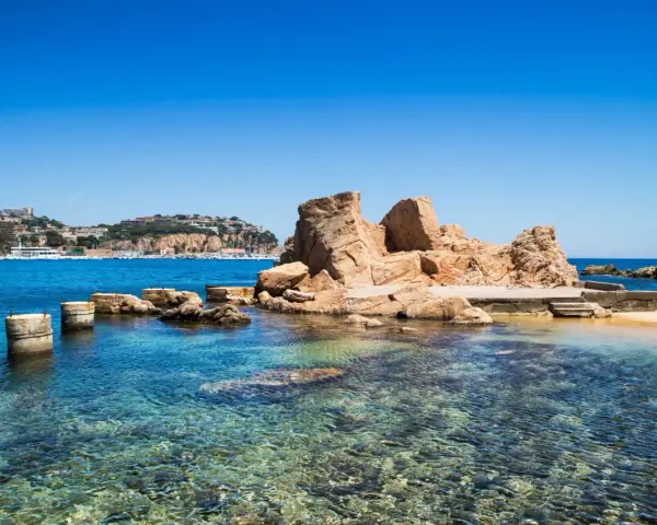 Un paseo marítimo en Sant Feliu de Guíxols, con rocas doradas y aguas cristalinas. El pueblo se alza en la colina.