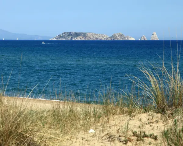 Dunas de hierba seca en primer plano, mar azul y un archipiélago rocoso en la distancia, evocando la belleza de la playa mas Pinell.