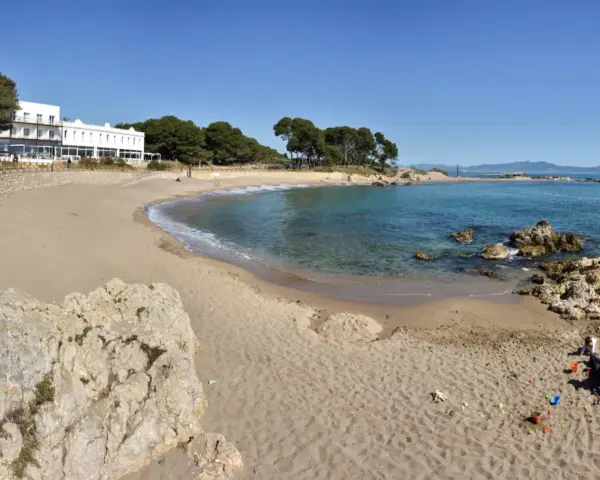 Platja Cala de la Creu: arena dorada, aguas turquesas y un hotel blanco con vistas al mar. Familias disfrutan del sol y juegan en la arena.