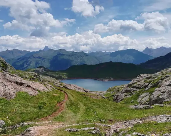 Sendero de montaña que conduce al sereno ibon de Estanes, rodeado de picos escarpados y un cielo azul con nubes algodonosas.