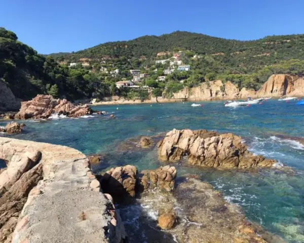 Un puente de piedra cruza aguas turquesas en la cala Canyet. Barcas flotan tranquilas mientras colinas verdes y casas blancas coronan el paisaje.
