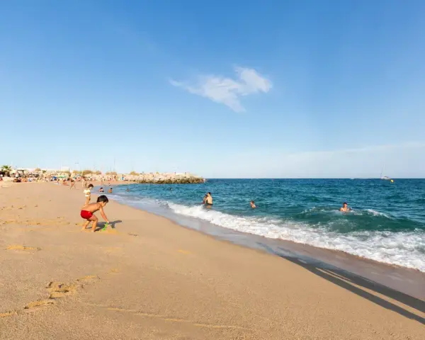 Un niño juega en la arena dorada de la playa de les barques, mientras otros disfrutan del mar azul y un velero navega en el horizonte.