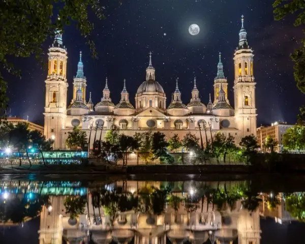 La majestuosa basílica del Pilar, iluminada bajo un cielo estrellado y la luna, se refleja en el río.