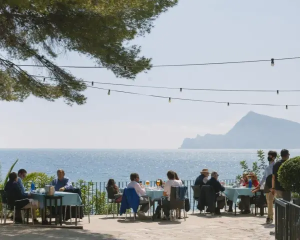 Gente disfrutando de una comida al aire libre con vistas al mar. Un lugar ideal donde comer en Altea, con mesas azules y un ambiente relajado bajo un árbol.