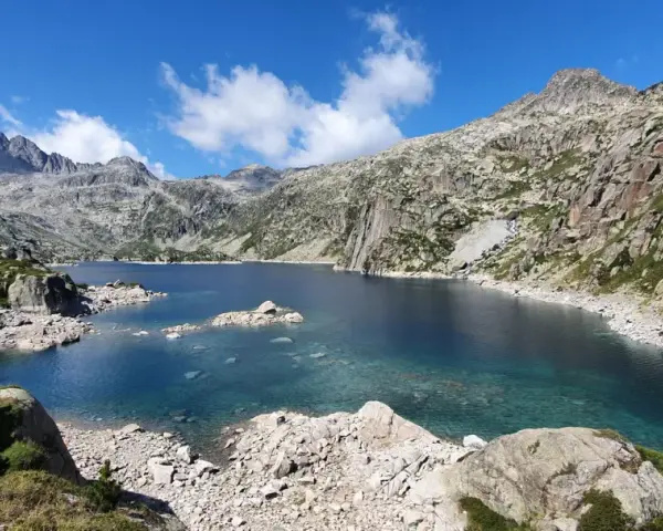 Lago de aguas cristalinas rodeado de montañas rocosas en el Vall de Boí. El cielo azul con nubes blancas contrasta con el verde de la vegetación.