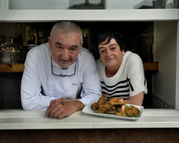 Chef y camarera sonríen tras ventana con plato de setas. Descubre dónde comer en Castro Urdiales.