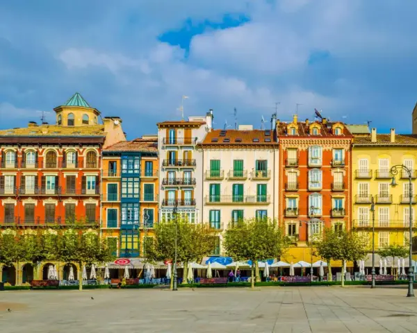 Fachada de edificios coloridos en una plaza, ideal para saber **qué ver en Pamplona**. Terrazas y árboles verdes bajo un cielo azul.