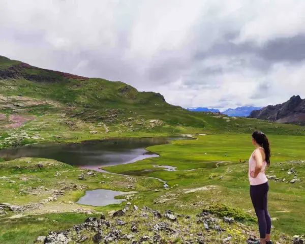 Mujer contempla los ibones del Valle de Tena, un paisaje de lagos y prados verdes bajo un cielo nublado.