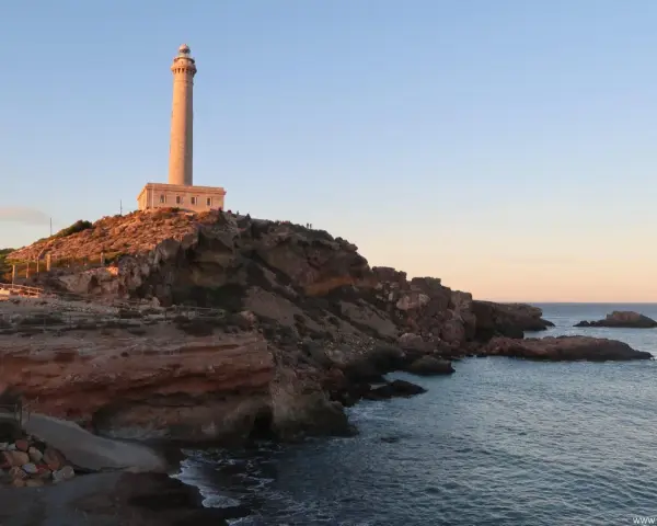 Faro sobre acantilados rojizos, con el mar azul y un cielo despejado. Un camino desciende hacia una pequeña cala roja.