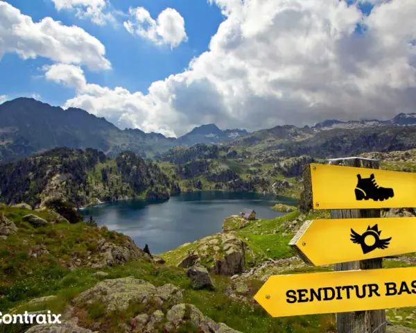 Paisaje de montaña con un lago azul, señalización de sendero y el letrero "SENDITUR BASIC", parte de la ruta de los 7 lagos Pirineos.