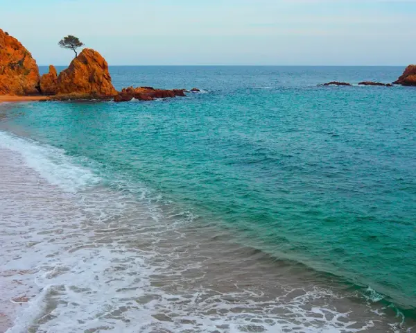 La platja mar menuda, con sus rocas anaranjadas y aguas turquesas, invita a soñar. Una ola rompe suavemente en la arena dorada.
