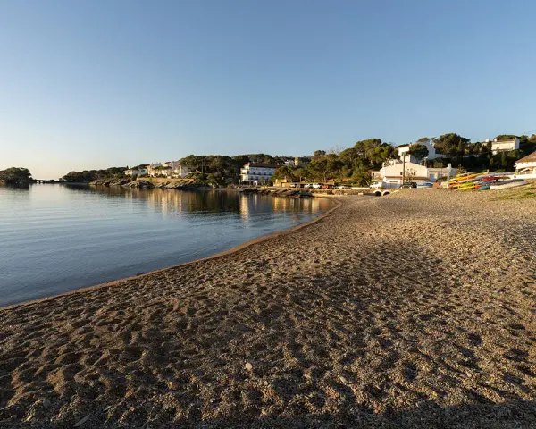 La platja gran de Cadaqués, con casas blancas y kayaks coloridos, se baña en la calma del mar al atardecer.