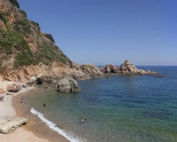 Les platges de Blanes ofereixen aigües cristal·lines i roques imponents sota un cel blau. Gent gaudeix del sol i del mar.