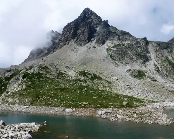 Paisaje montañoso con un lago de aguas tranquilas en primer plano. Las laderas están cubiertas de rocas y vegetación verde, con picos escarpados y nubes en el cielo. Los **ibones de Arriel** reflejan la majestuosidad del entorno.