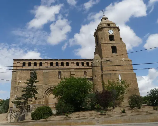 Iglesia de piedra con torre y reloj, rodeada de vegetación. Un día soleado en Alberuela de Tubo.