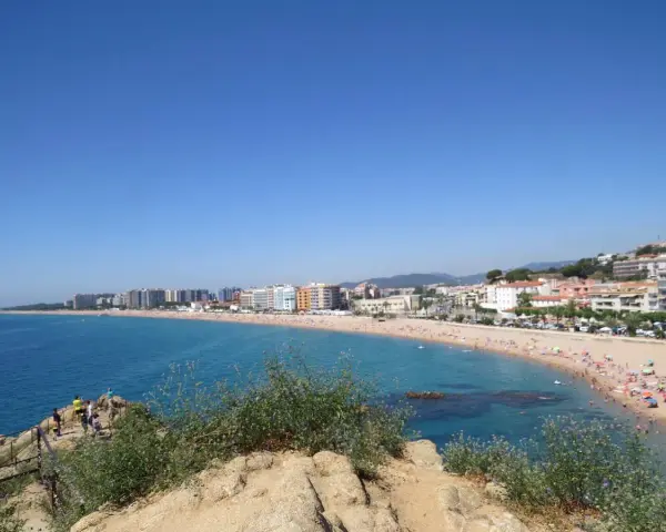 Vista panorámica de la playa S'Abanell en Blanes, con el mar azul intenso, arena dorada repleta de gente y edificios costeros bajo un cielo despejado.