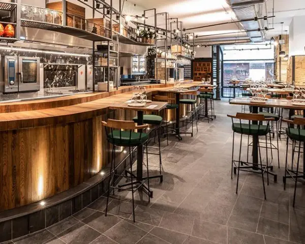 Bar moderno con taburetes verdes y mesas de madera. Un lugar ideal donde comer en Salamanca, con cocina a la vista y ambiente acogedor.