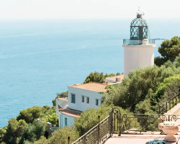 El faro de Sant Sebastià se alza sobre el mar azul, rodeado de vegetación exuberante.