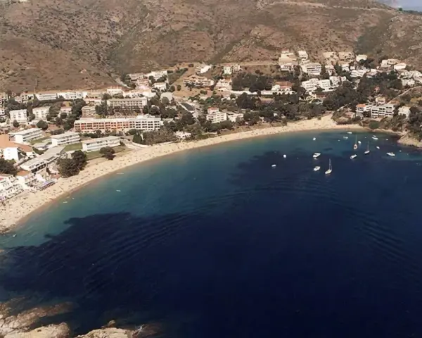Vista aérea de la playa Almadrava en Roses, con edificios blancos y barcos anclados en aguas azules.