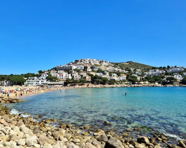 Vista de la platja de Montgó, con gente disfrutando del sol y el mar turquesa, y casas blancas escalonadas en la colina.