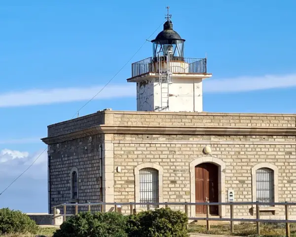 Faro de piedra con torre blanca y cúpula negra, vigilando el mar azul cerca del far port de la selva.