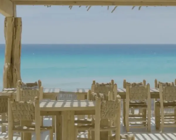 Mesas y sillas de madera rústica en la terraza de restaurantes en Formentera, con el mar turquesa de fondo.