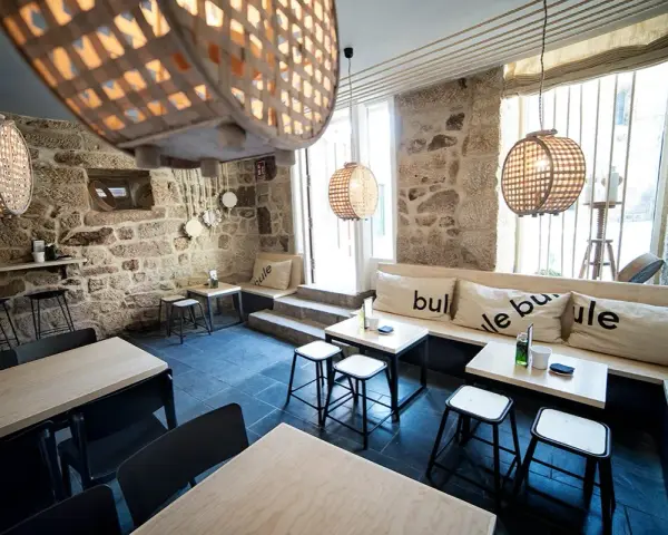 Ambiente acogedor para comer en Allariz. Mesas de madera, bancos con cojines y lámparas de mimbre en un local de piedra.