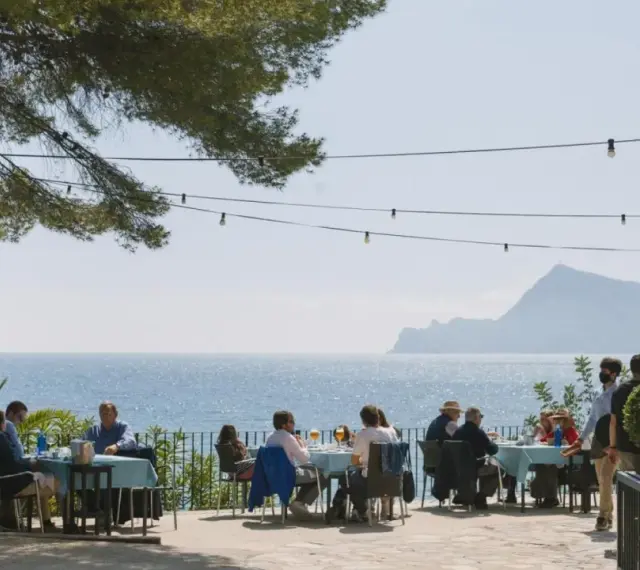 Gente disfrutando de una comida al aire libre con vistas al mar. Un lugar ideal donde comer en Altea, con mesas azules y un ambiente relajado bajo un árbol.