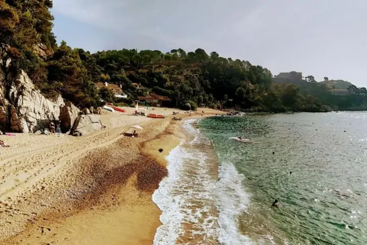 Playa de Santa Maria de Llorell con rocas, pinos y gente disfrutando del sol y el mar.