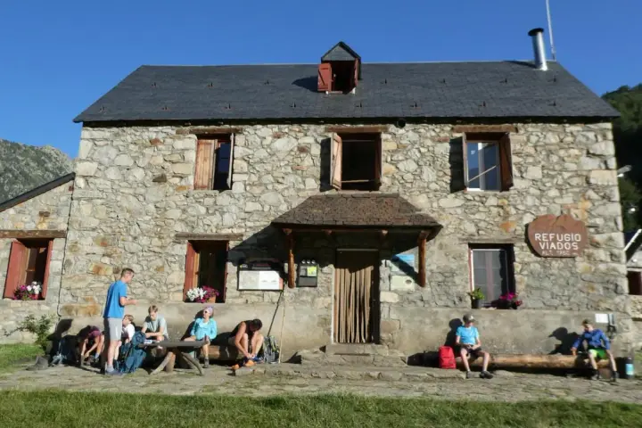 Grupo de excursionistas descansa frente al Refugio Viados, parte de la ruta de los tres refugios. Montañas y cielo azul al fondo.