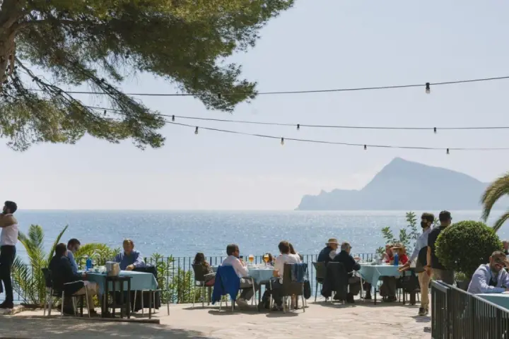 Gente disfrutando de una comida al aire libre con vistas al mar. Un lugar ideal donde comer en Altea, con mesas azules y un ambiente relajado bajo un árbol.