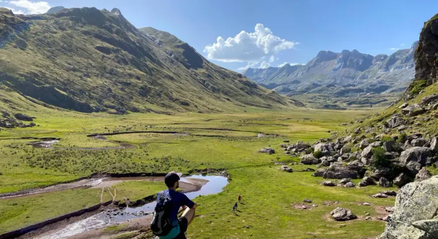 Un excursionista contempla un vasto valle de hecho, con un río serpenteante y montañas majestuosas bajo un cielo azul.