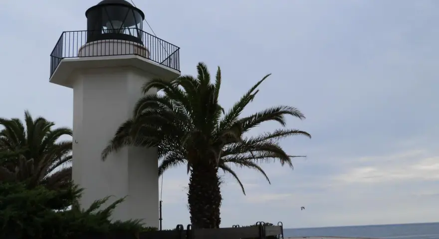 El faro de Palamós, blanco y esbelto, se alza junto a palmeras y el mar azul.