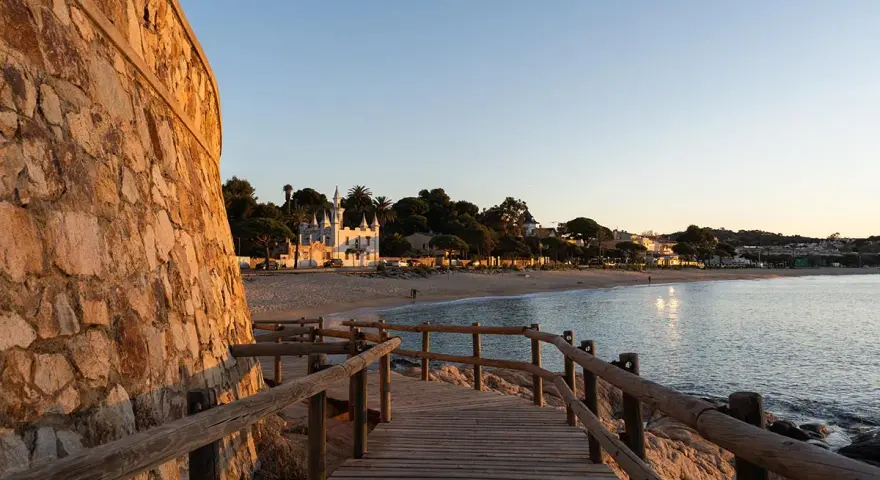 Pasarela de madera hacia la platja Sant Pol, Sant Feliu de Guíxols. Edificios blancos y palmeras bordean la arena dorada bajo un cielo azul.