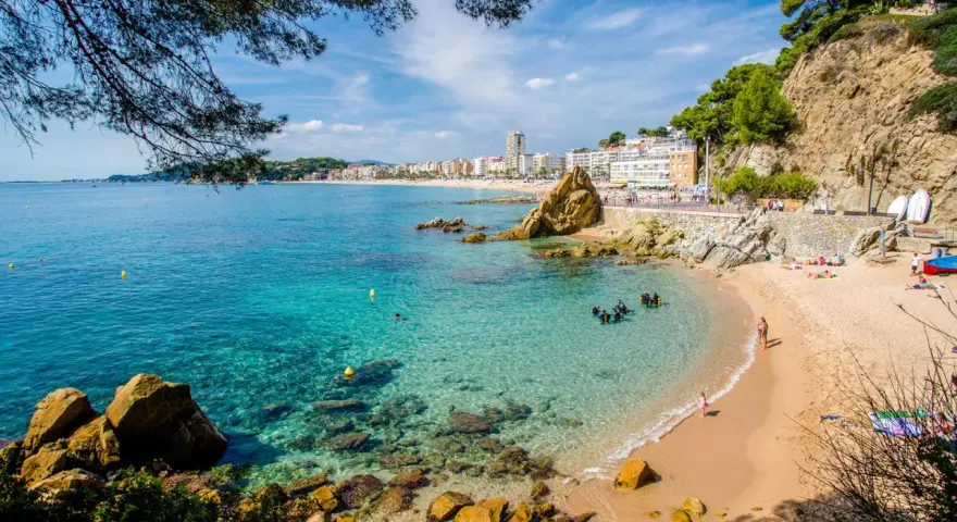 Una caleta en la Costa Brava con aguas cristalinas, buceadores explorando el fondo rocoso y una playa con gente disfrutando del sol.