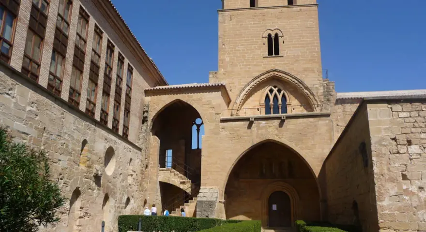 El **castillo de Alcañiz**, con su imponente torre y arcos, se alza bajo un cielo azul. Jardines cuidados y muros de piedra evocan su historia.