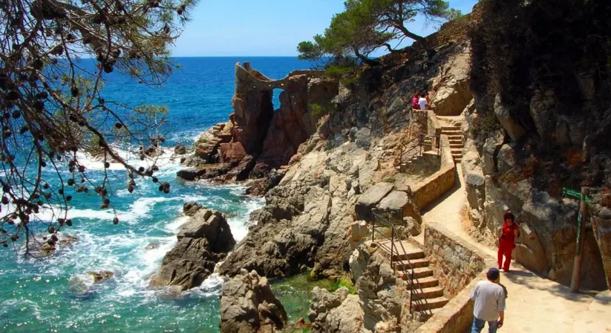 Un camino de ronda con escaleras de piedra serpentea por la costa rocosa. El mar turquesa rompe contra las rocas.