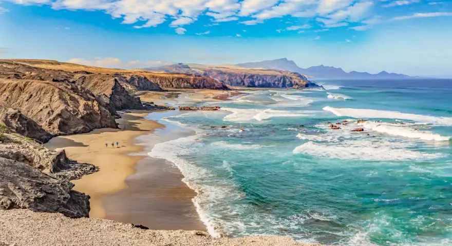 Paisaje de las playas de Fuerteventura con acantilados rocosos, arena dorada y el mar turquesa con olas rompiendo.